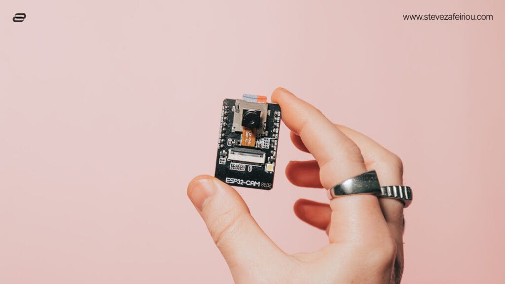 Hand holding a compact ESP32-CAM development board featuring a camera module against a pastel pink background.