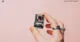 Hand holding a compact ESP32-CAM development board featuring a camera module against a pastel pink background.