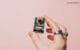 Hand holding a compact ESP32-CAM development board featuring a camera module against a pastel pink background.